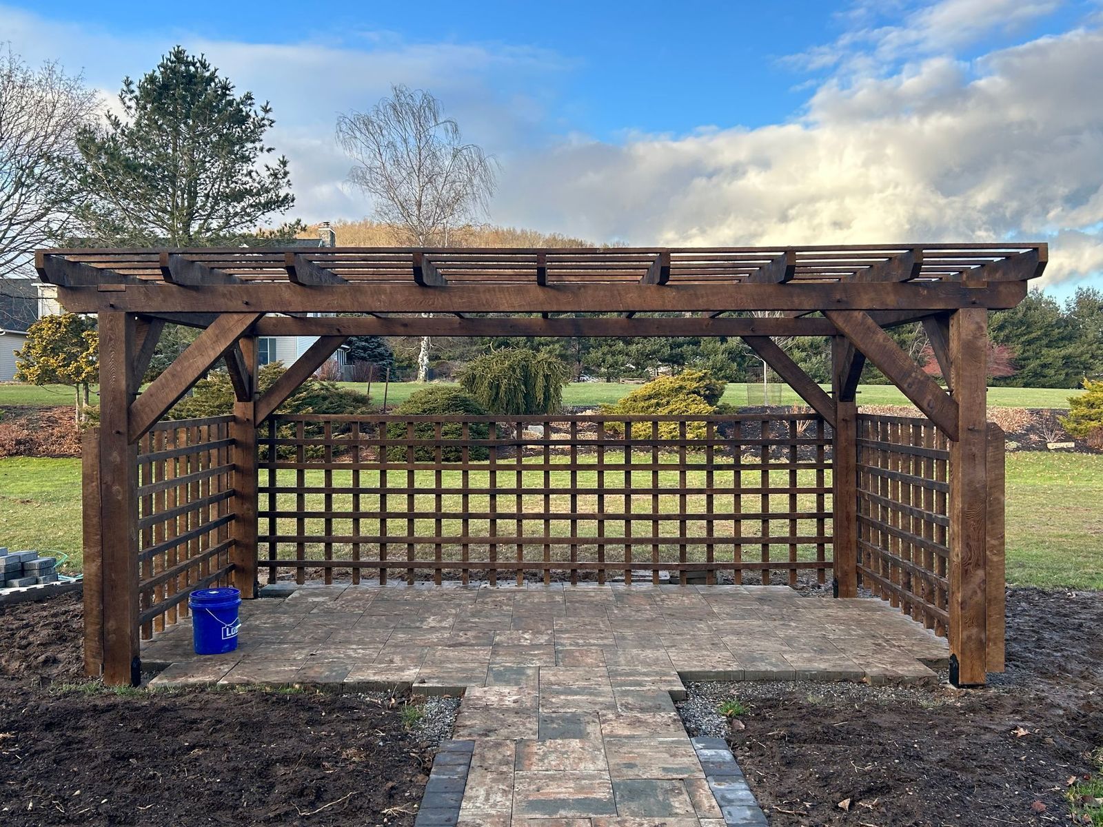 A brown wooden pergola with a lattice back and side wall sits on a paved patio in a grassy, landscaped yard.