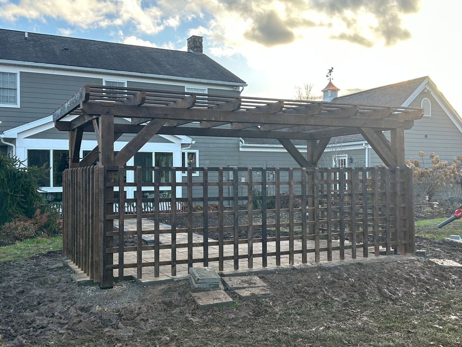 A dark-stained wooden pergola by CTFS with a lattice structure stands in a backyard in front of a house. West Virginia. 