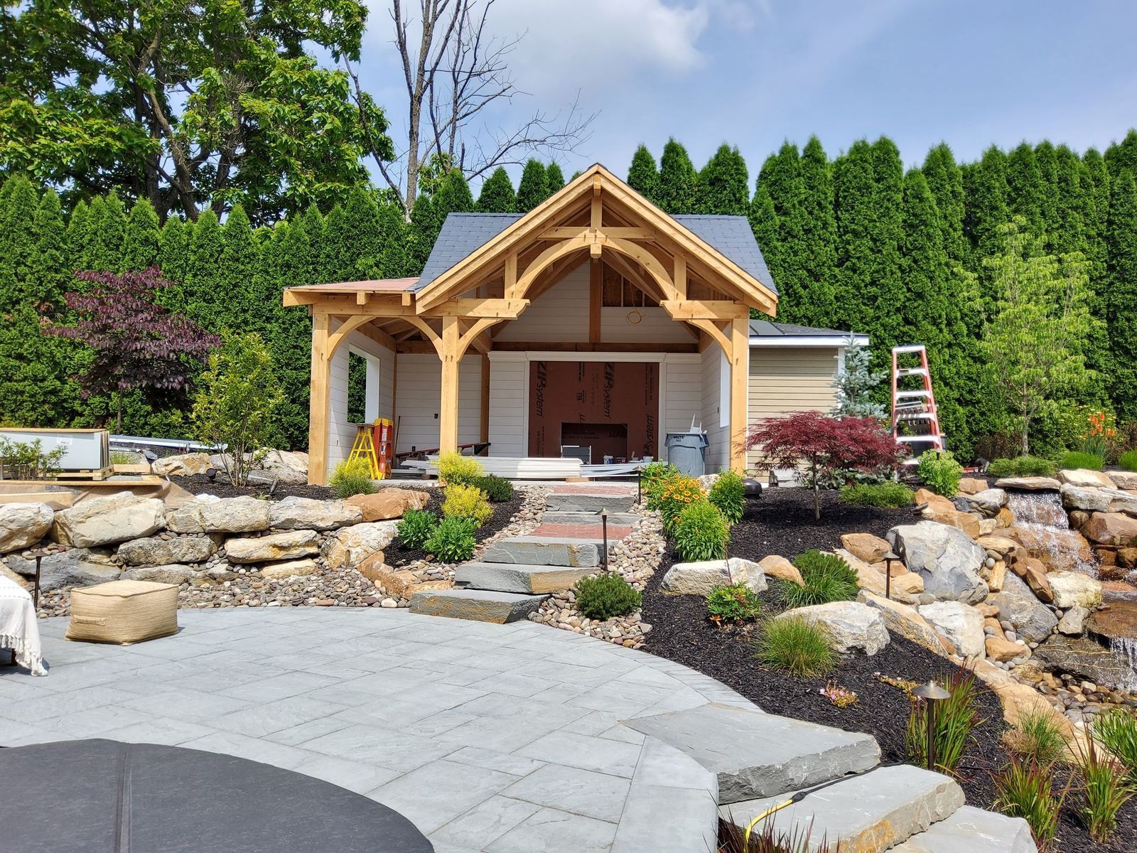 An outdoor patio featuring a timber-frame pavilion with a stone fireplace, set among landscaped rocks and green hedges.