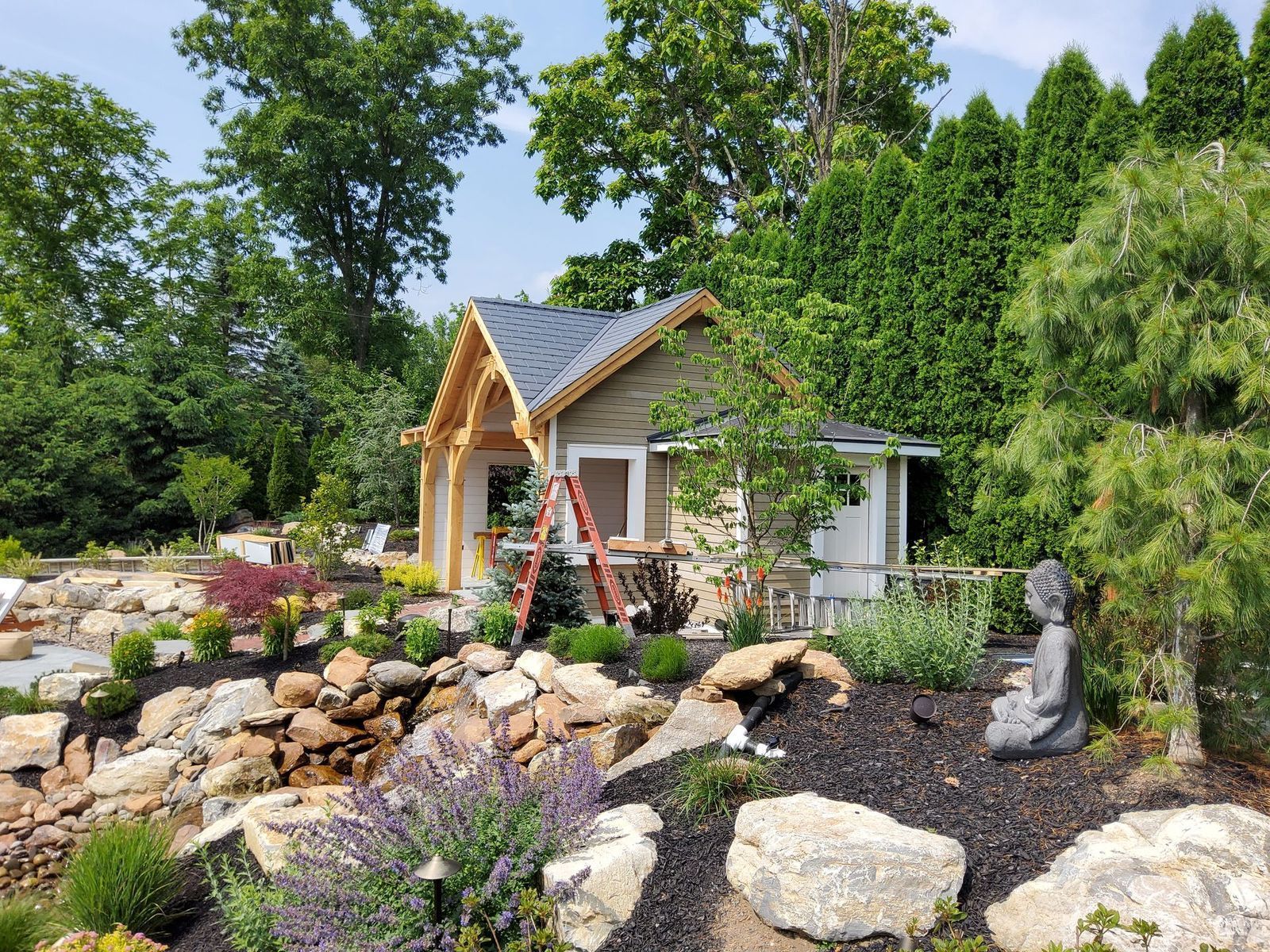 A small, shingled shed with decorative wooden trim sits in a landscaped garden with a rock pond and a Buddha statue.