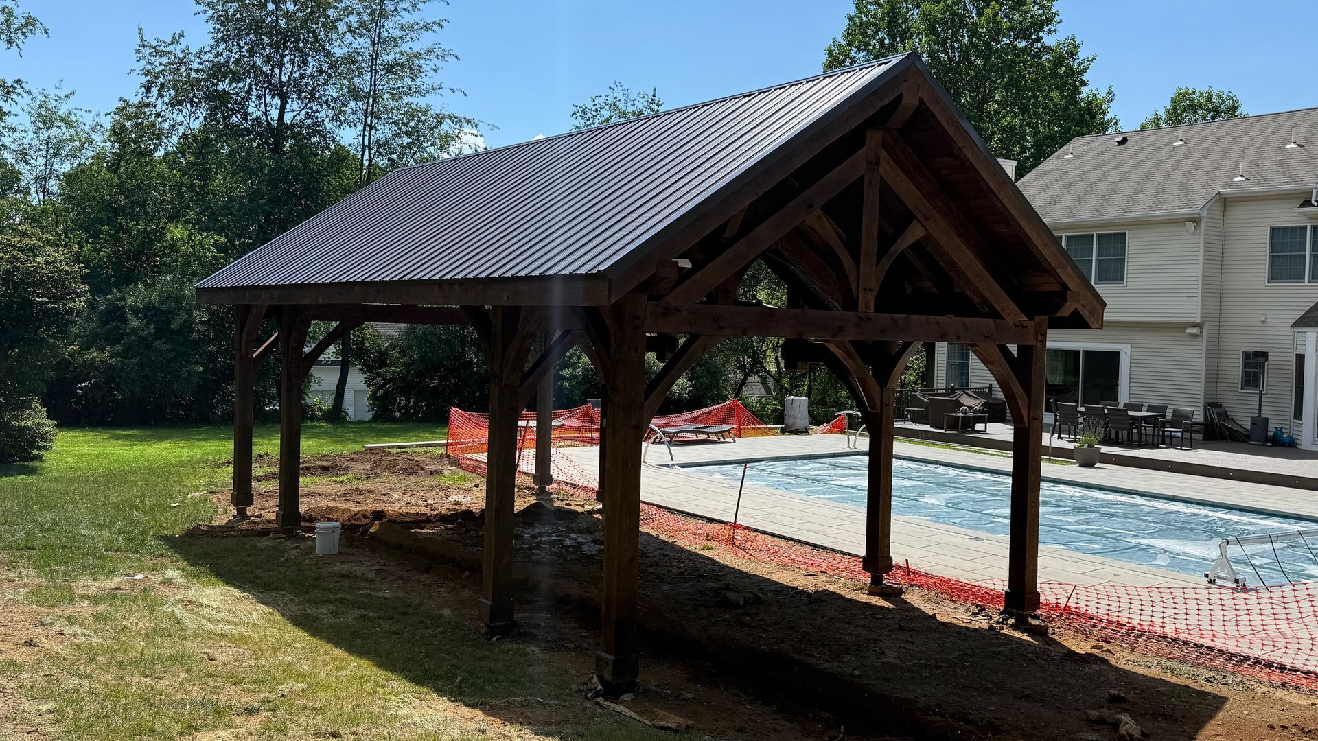 A large, dark wood pavilion structure with a metal roof stands in a backyard near a swimming pool and house.