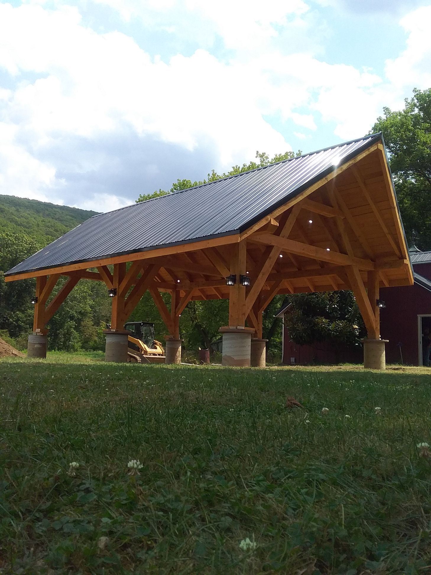 A large, wooden, open-air pavilion with a dark gabled roof set on concrete pillars in a grassy field with trees.