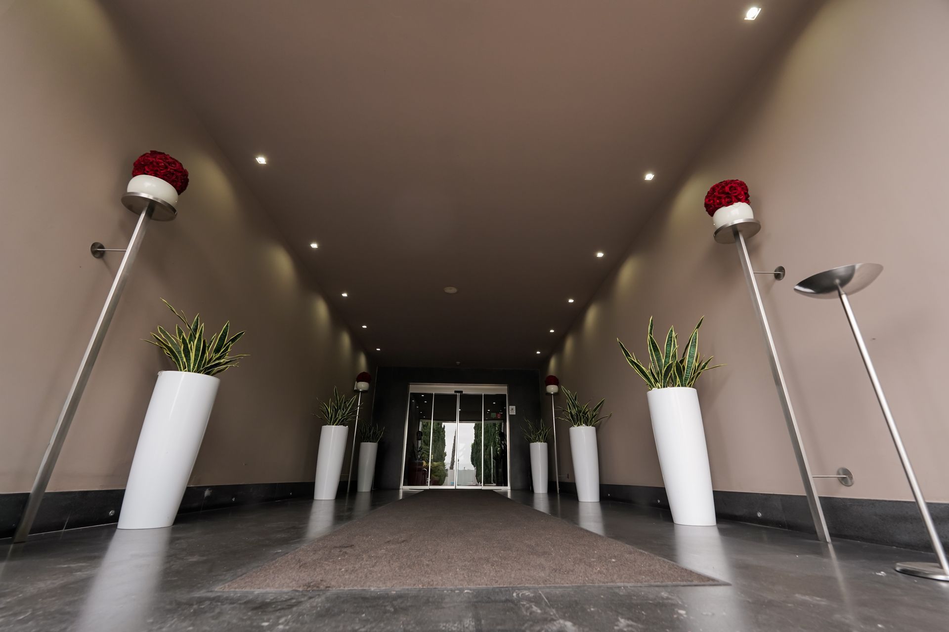 Ingresso di un edificio moderno con alte fioriere bianche, cappelli di Babbo Natale rossi e una porta a vetri in fondo a un corridoio.