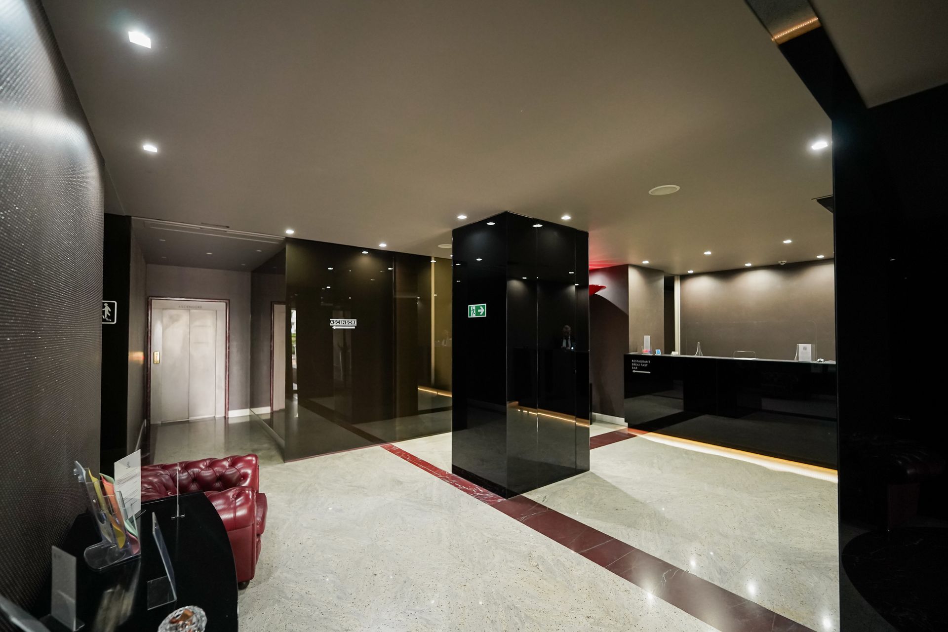 Hall d'albergo moderna con colonne nere lucide, sedute rosse e luci calde sul soffitto.
