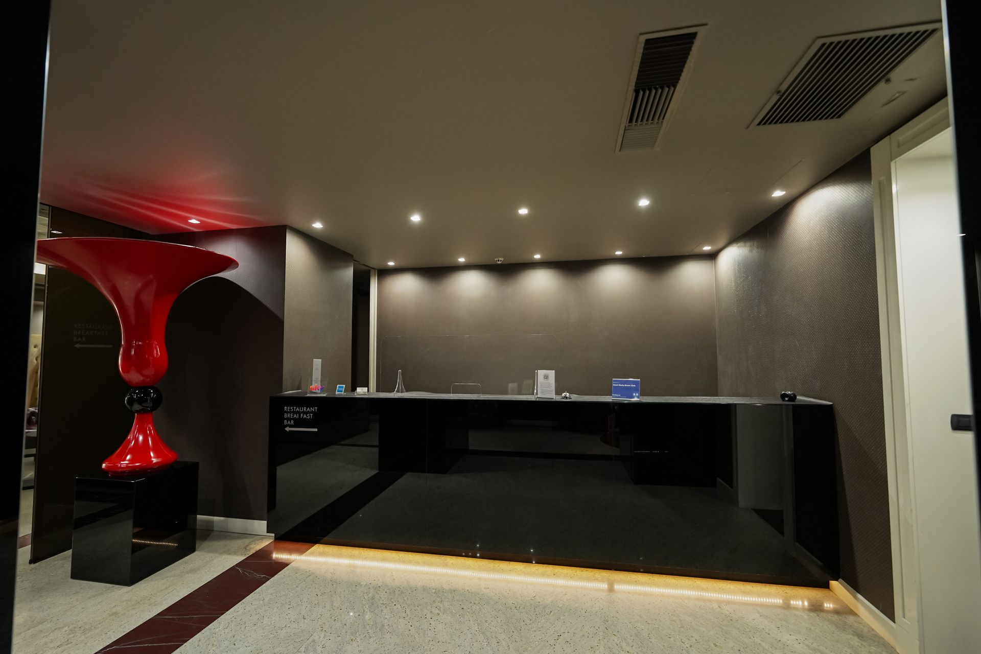 Banco reception moderno nella hall con piano nero, decorazioni scultoree rosse e faretti a incasso sul soffitto.