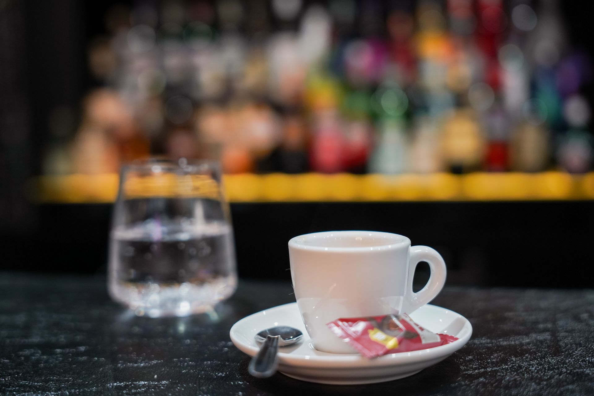 Una tazza da caffè bianca su un piattino con bustina di zucchero e cucchiaino sul bancone del bar, un bicchiere d'acqua accanto.