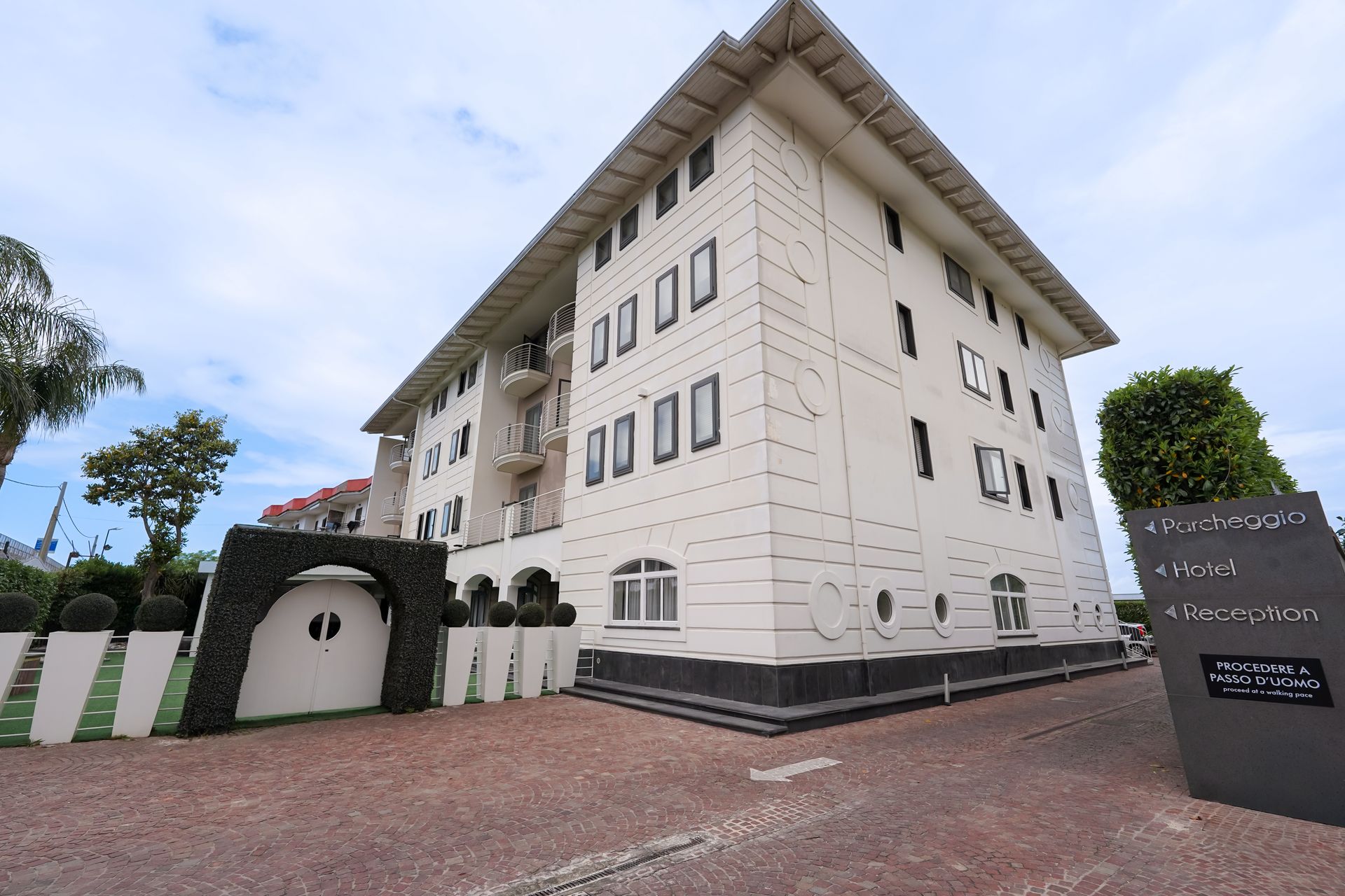 Edificio bianco a più piani con ingresso ad arco e cortile pavimentato sotto un cielo nuvoloso