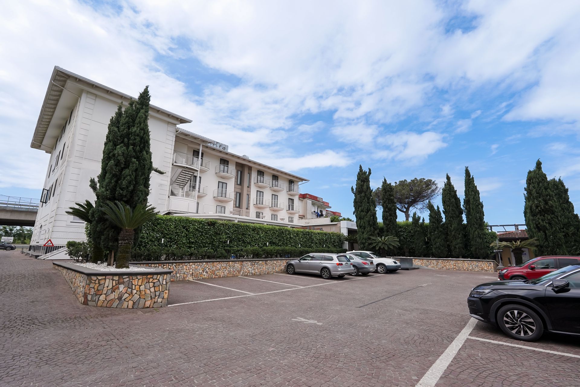 Grande edificio bianco con alti cipressi e auto parcheggiate in un piazzale ghiaioso sotto un cielo parzialmente nuvoloso