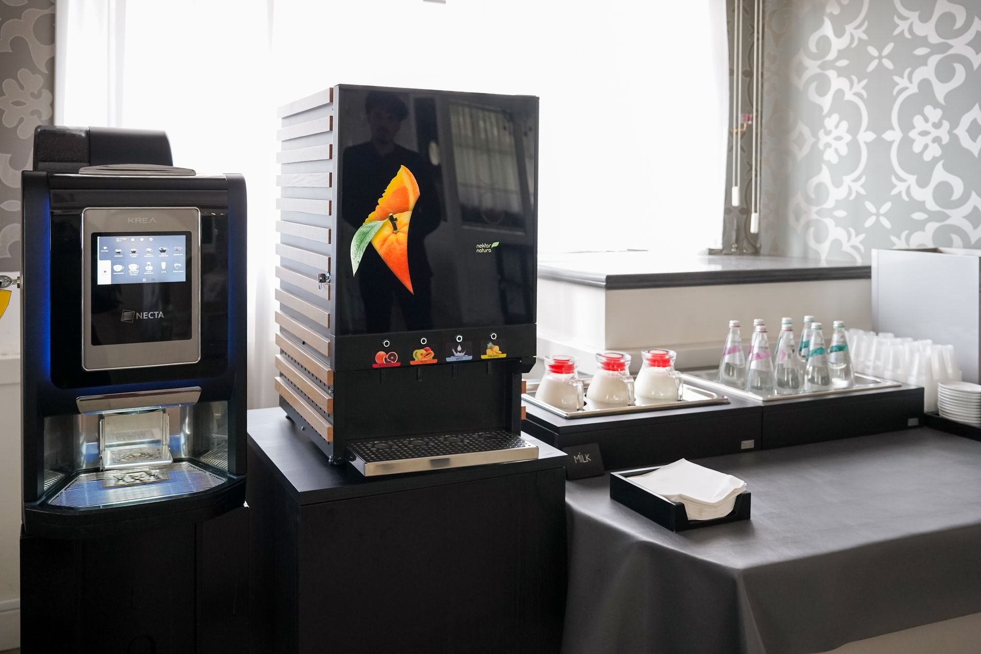 Postazione per caffè e bevande con due macchine, tazze e condimenti su un bancone nero.