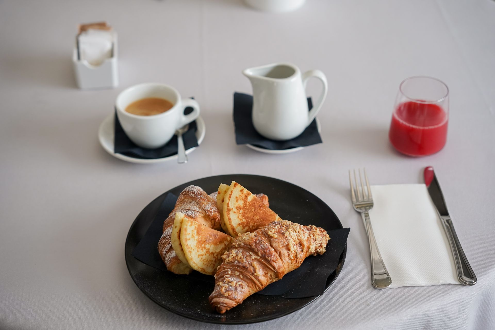 Piatto per la colazione con croissant e patate, caffè, lattiera, succo di frutta e posate su un tavolo bianco.
