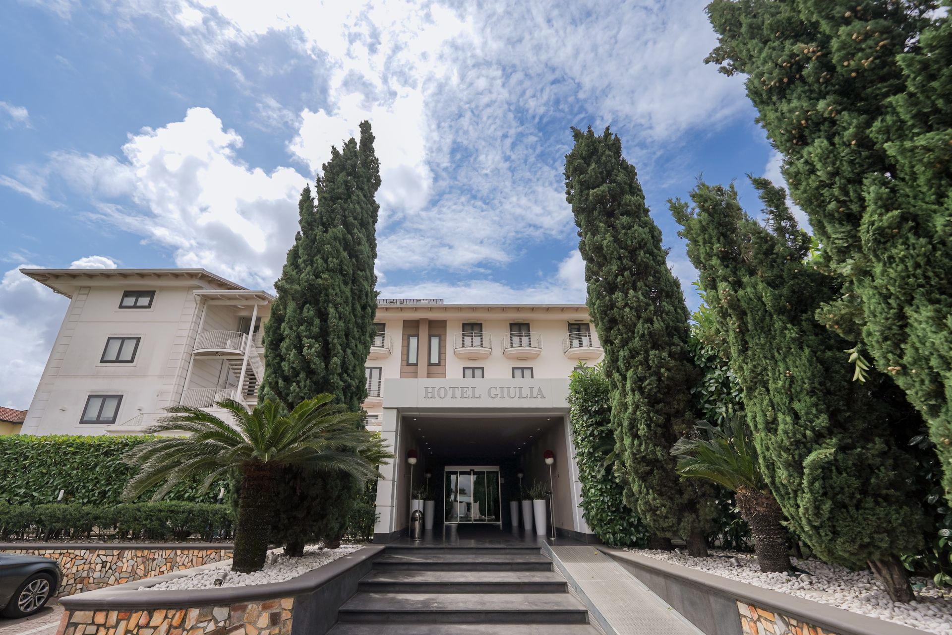 L'ingresso dell'edificio è incorniciato da alti cipressi sotto un cielo azzurro nuvoloso.