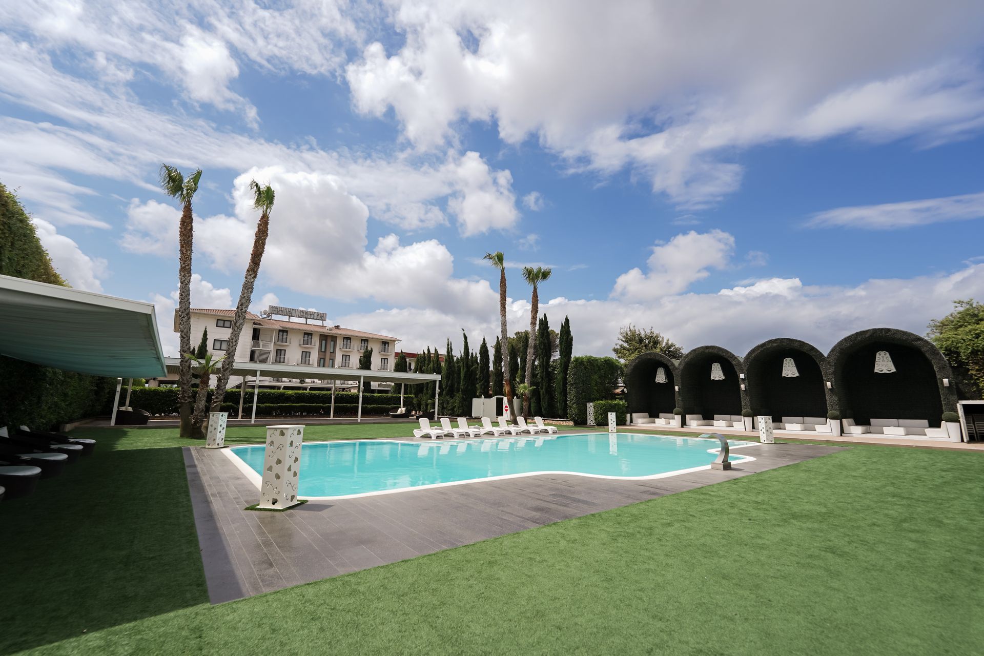 Piscina all'aperto con acqua turchese, palme e gazebo neri ombreggiati sotto un cielo parzialmente nuvoloso.