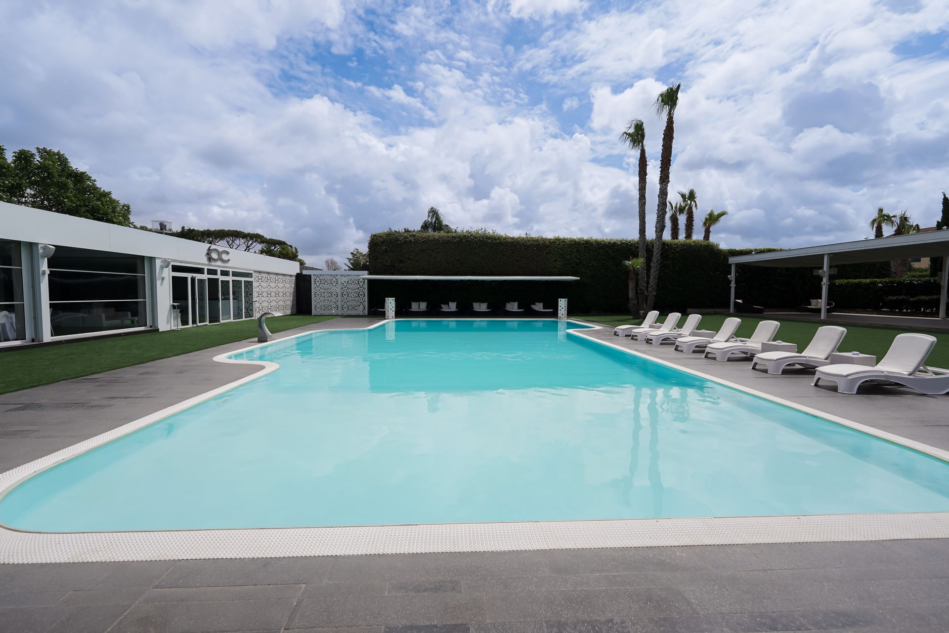 Piscina all'aperto con acqua turchese, lettini bianchi e un cielo nuvoloso.