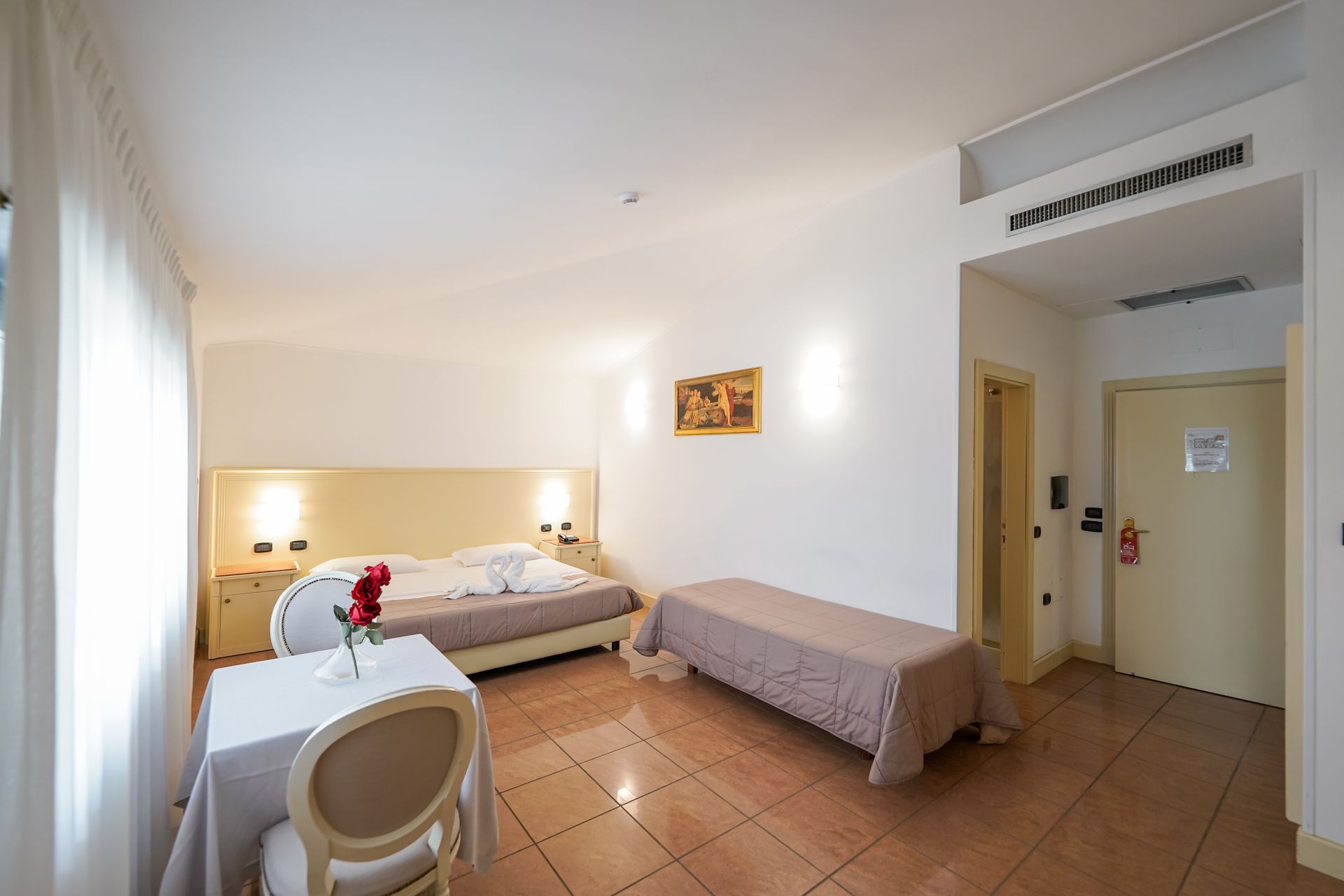 Piccola camera d'albergo con due letti, una sedia da scrivania, pavimento piastrellato e una porta aperta che dà su un corridoio.