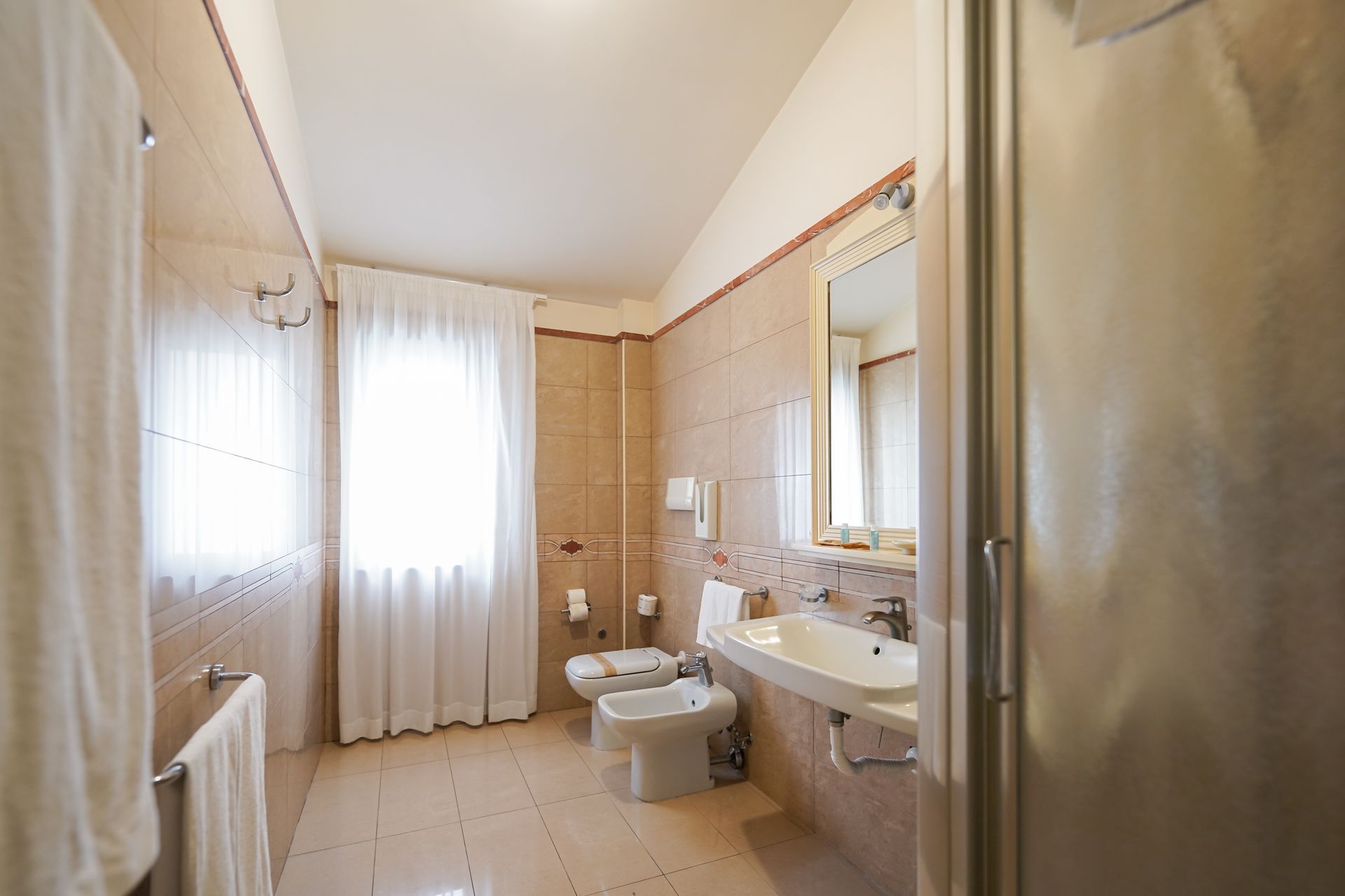 Piccolo bagno beige con lavabo, bidet, WC, box doccia e finestra con tende.