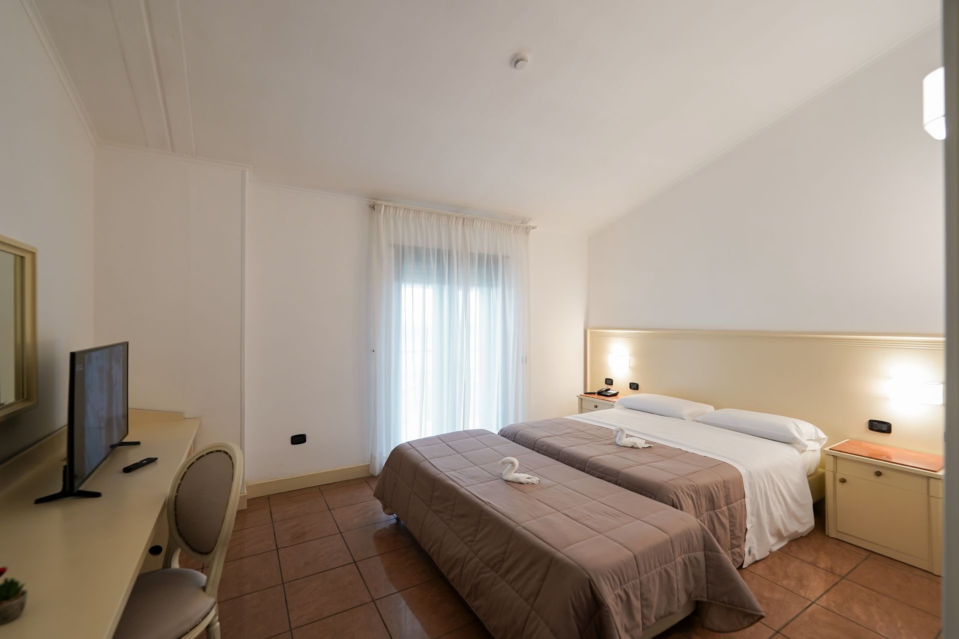 Camera da letto luminosa con letto, scrivania, TV, lampade da comodino e finestra con tende trasparenti.