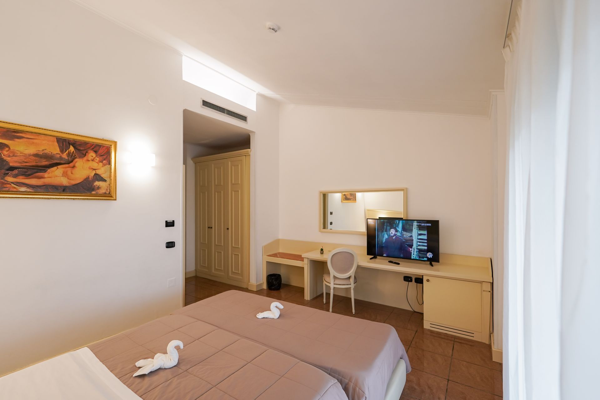 Camera da letto luminosa con letto, scrivania, sedia, specchio e TV su pareti bianche.