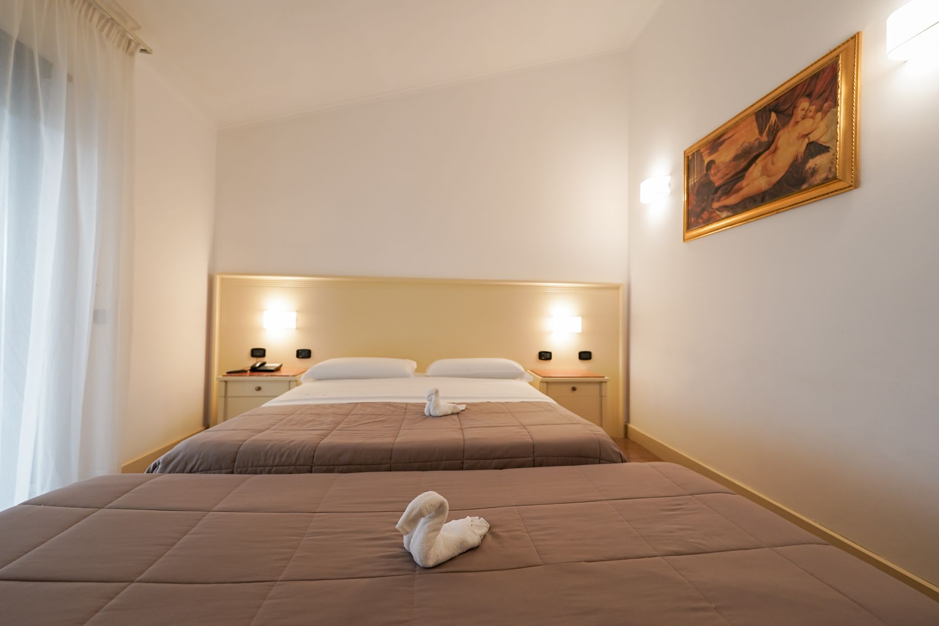 Due letti singoli ben rifatti in una luminosa camera d'albergo minimalista, con lenzuola beige e un quadro incorniciato alla parete.