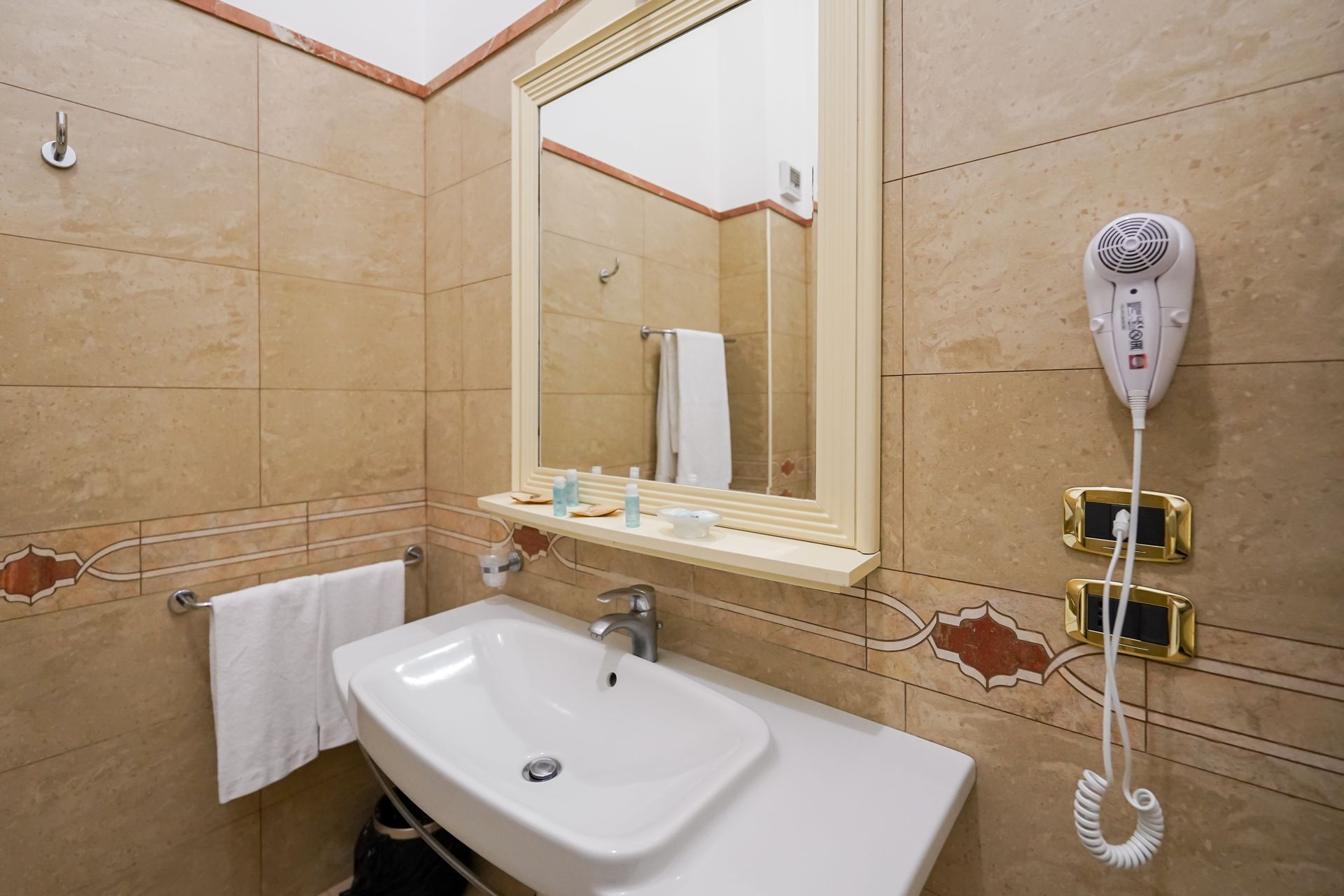 Piccolo bagno beige con lavandino, specchio, asciugamano e asciugacapelli a parete.