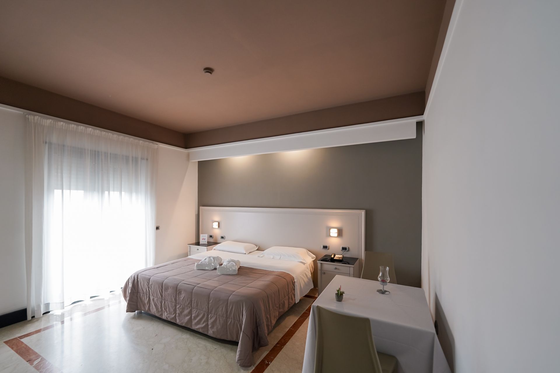 Camera da letto moderna con letto, comodini, pareti grigie e tende trasparenti accanto a una porta scorrevole in vetro.