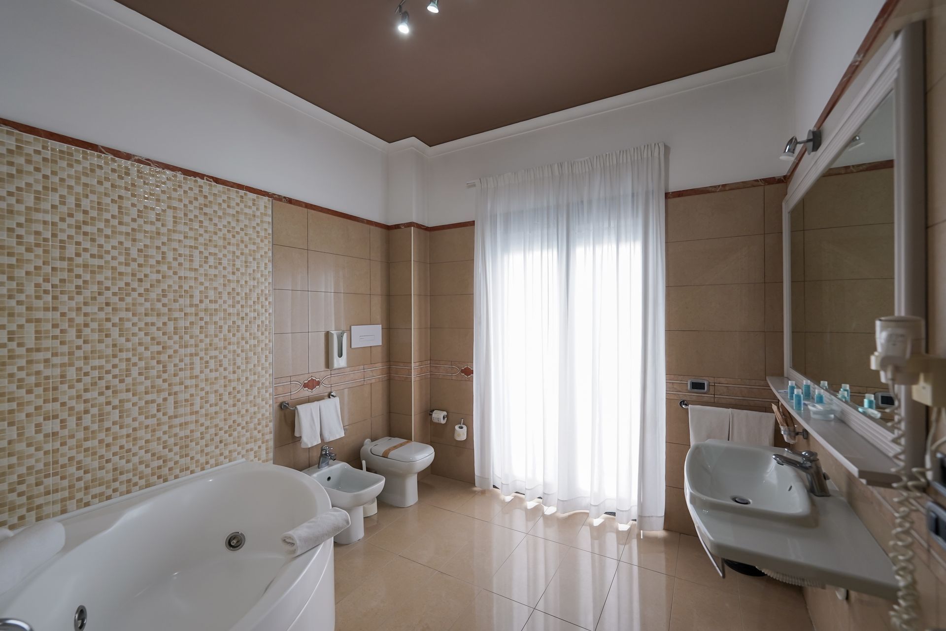 Bagno moderno con vasca da bagno, lavabo, WC e grande specchio accanto a una finestra luminosa con tende trasparenti.