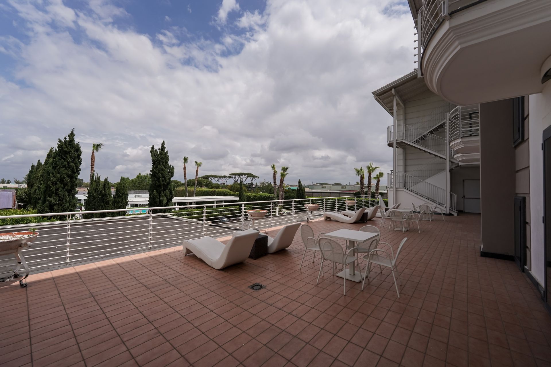 Terrazza con sedie a sdraio bianche, tavolo in vetro e vista sul cielo nuvoloso e sul giardino paesaggistico.