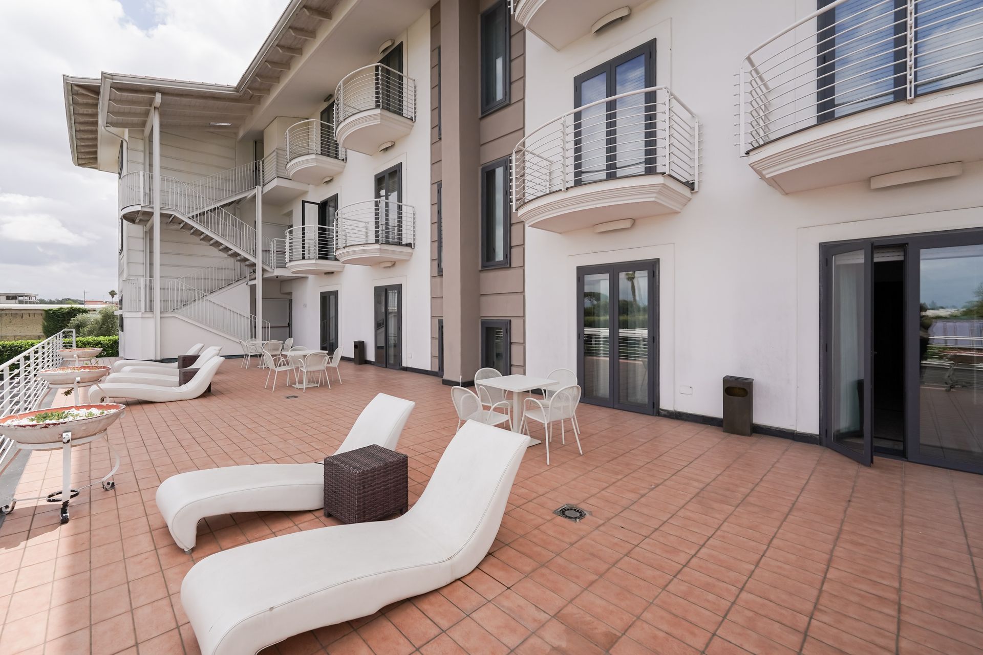 Ampia terrazza con lettini bianchi, piastrelle in mattoni e balconi degli appartamenti sotto una tettoia.