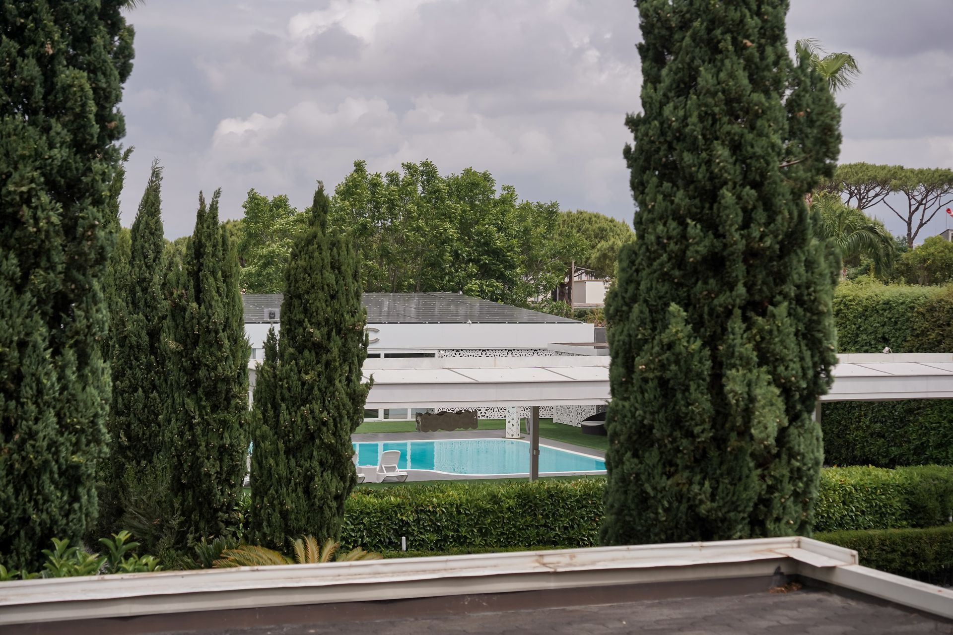Alti cipressi incorniciano una vista nuvolosa di una piscina e di un edificio moderno dietro una recinzione di legno.