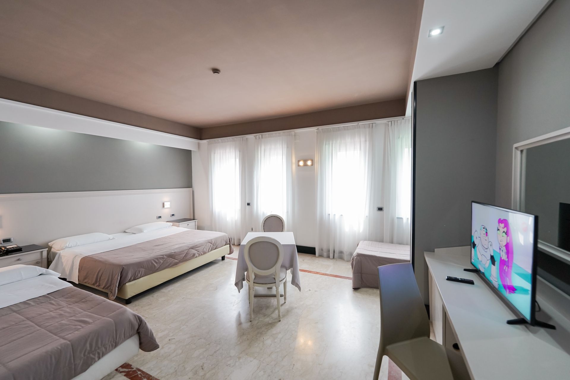 Luminosa camera d'albergo con due letti, una scrivania, TV e luce naturale che filtra attraverso tende trasparenti.