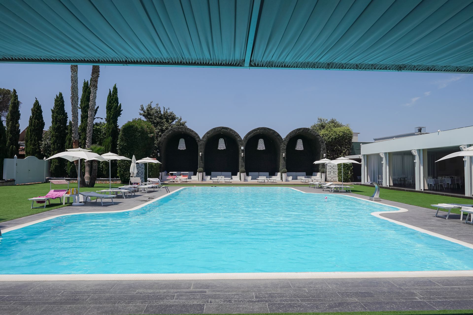 Piscina moderna del resort con acqua azzurra, lettini, ombrelloni e cabine ombreggiate sotto una tettoia a righe.