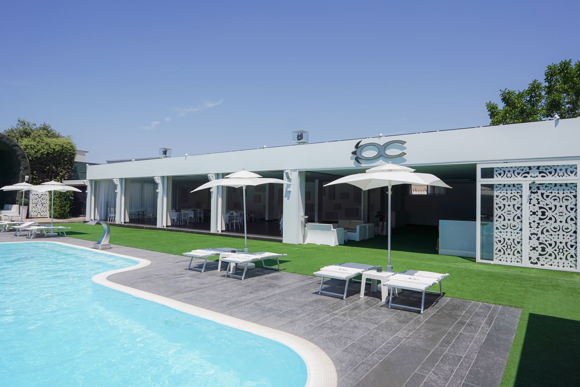 Area relax a bordo piscina con ombrelloni bianchi, lettini prendisole e un edificio moderno sotto un cielo azzurro limpido.