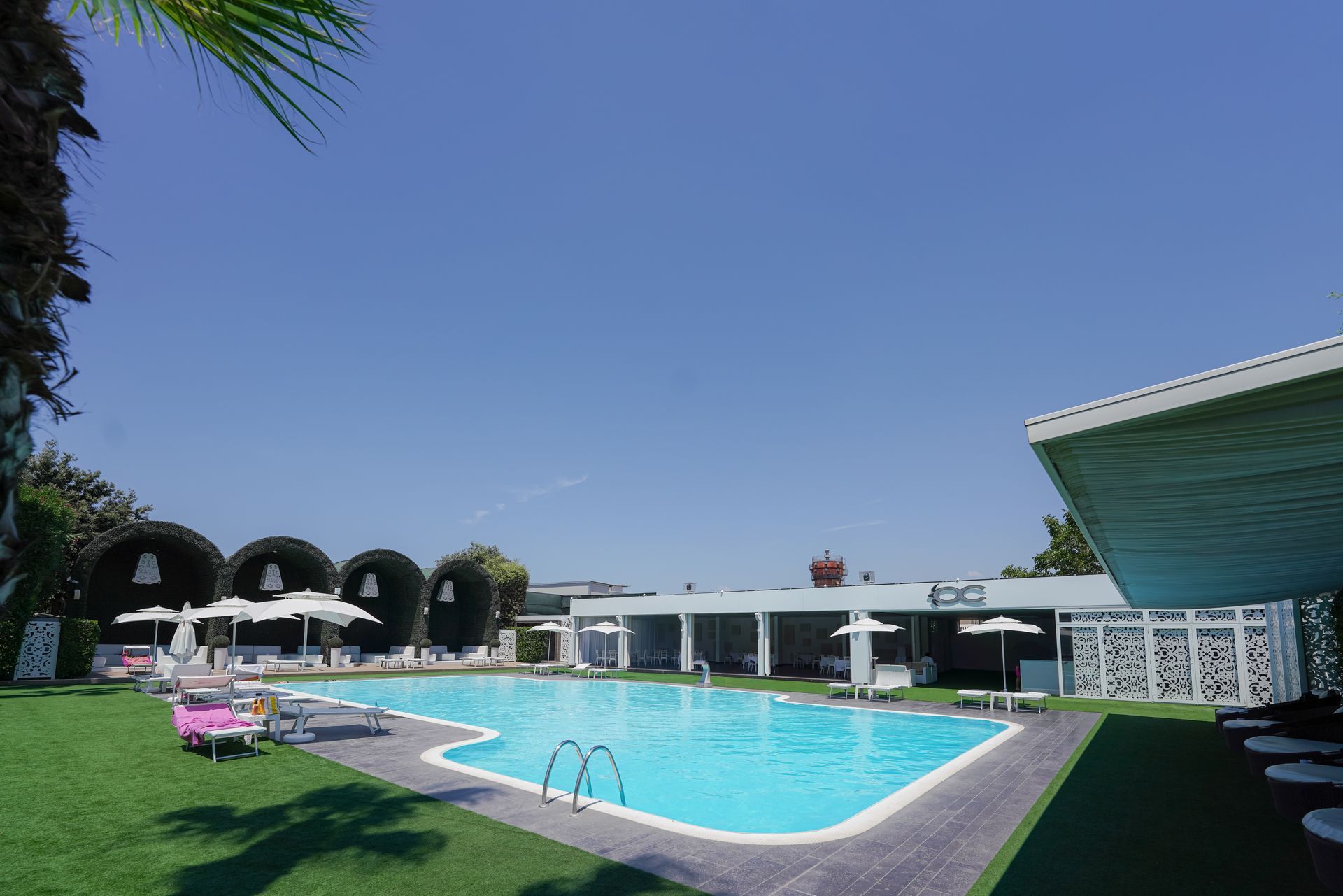 Piscina del resort con acqua azzurra, cabine bianche, prato verde e un cielo limpido in una giornata di sole.