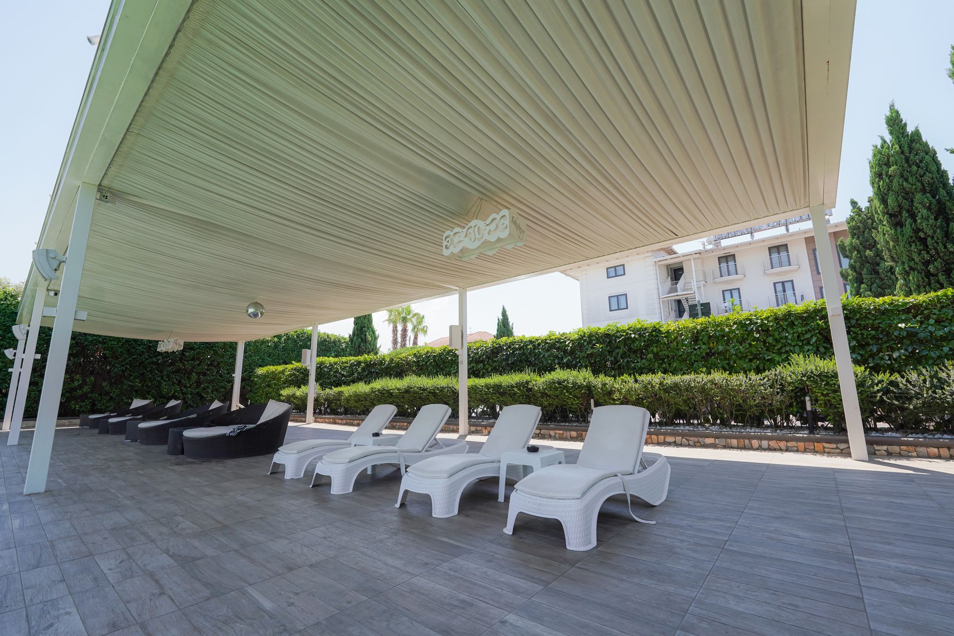Patio esterno con sedie a sdraio bianche sotto un grande gazebo a righe, siepi e palazzi sullo sfondo.