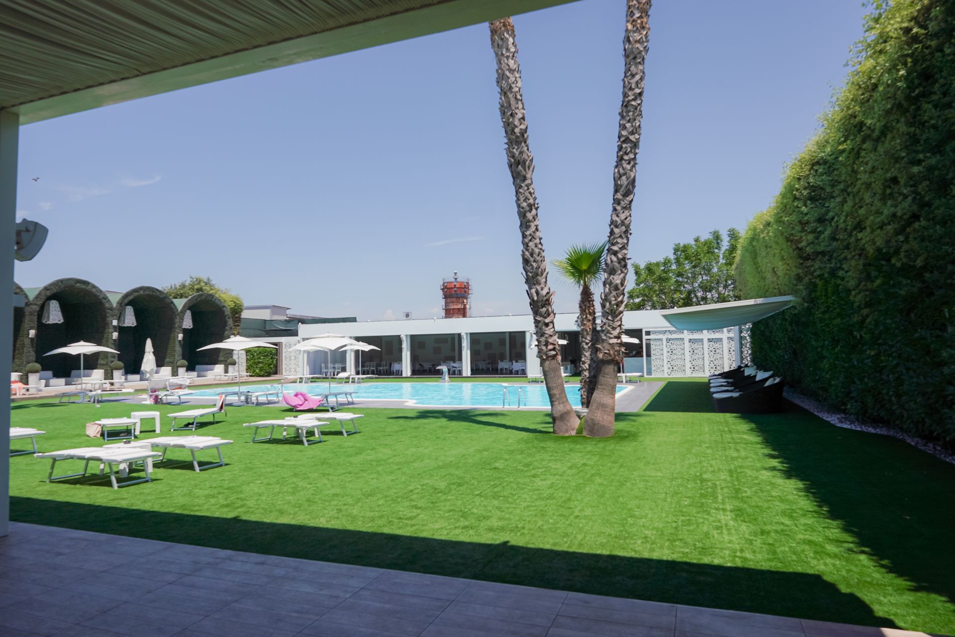 Giardino soleggiato con piscina, prato verde, sedie a sdraio e palme sotto un patio ombreggiato.