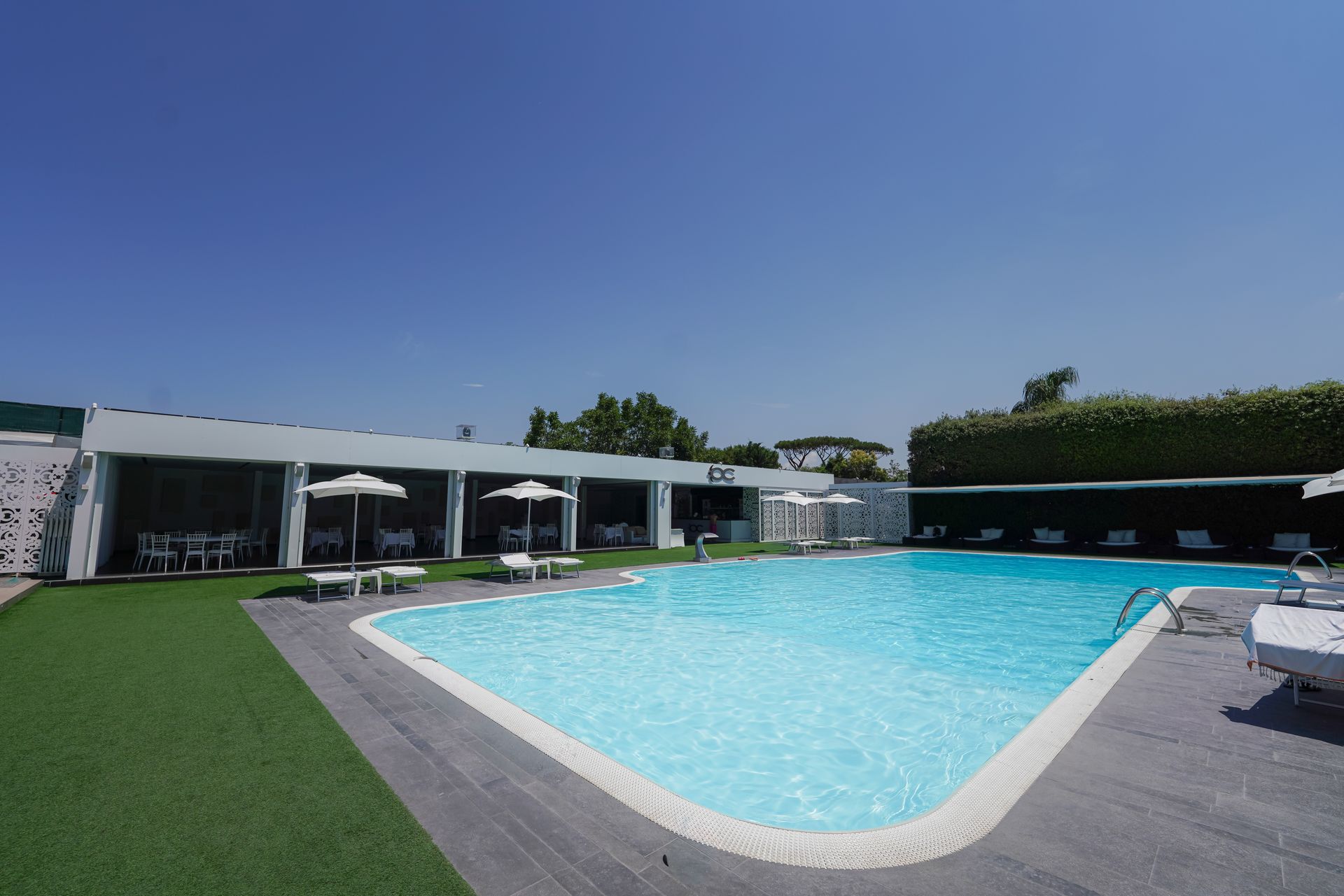 Piscina all'aperto accanto a un edificio moderno, con acqua azzurra, lettini bianchi e prato verde.