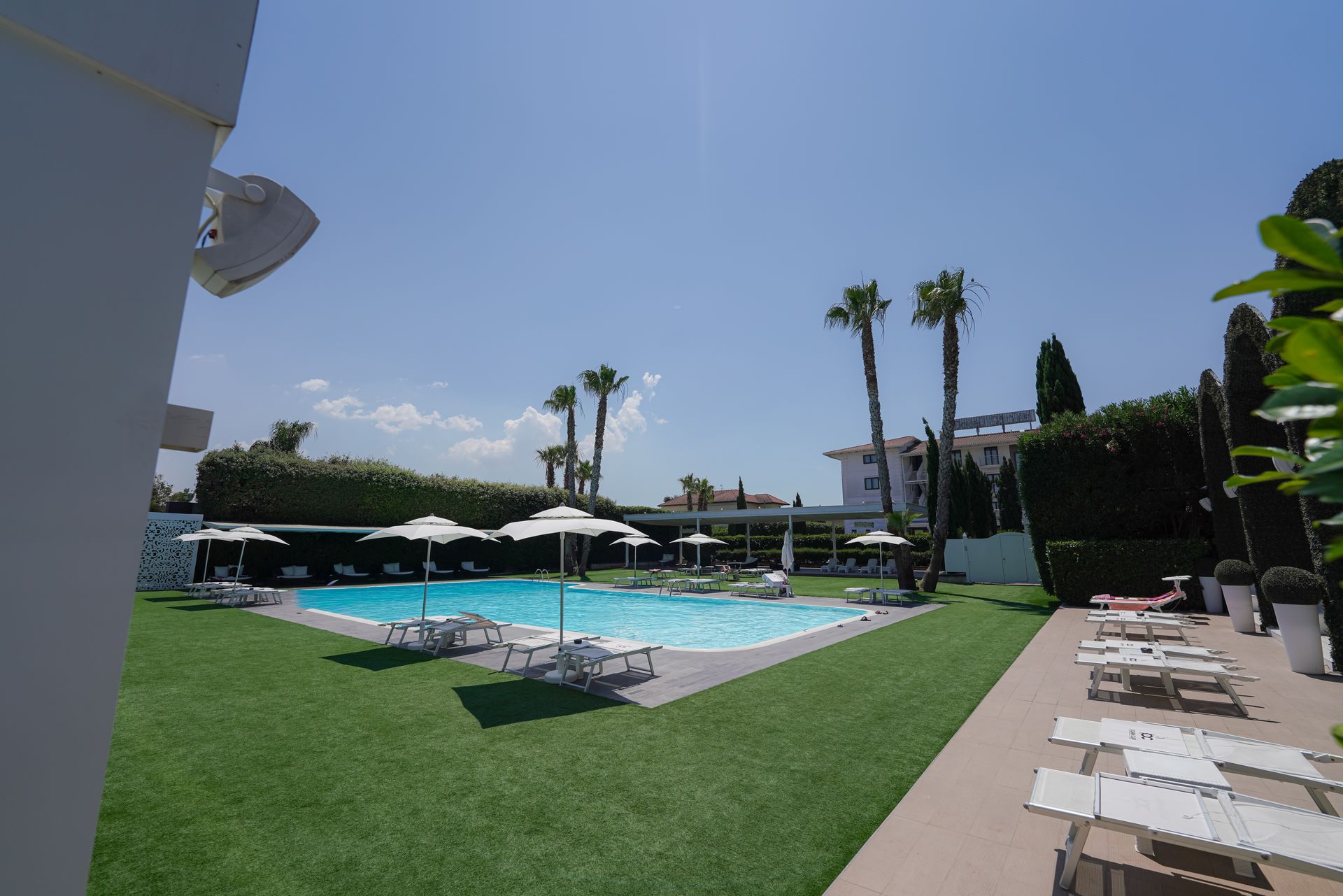 Piscina del resort con lettini, ombrelloni, palme e prato verde sotto un cielo azzurro brillante.