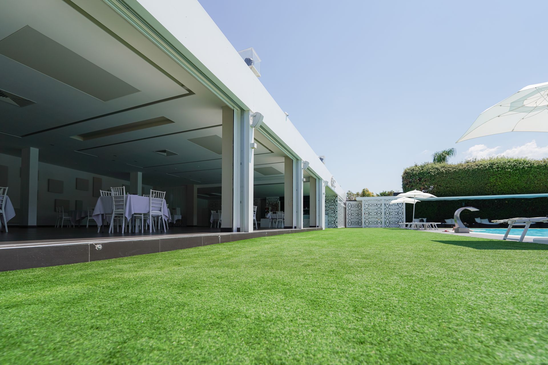 Patio moderno con area salotto coperta, prato verde e sedie a sdraio all'aperto sotto un cielo azzurro.