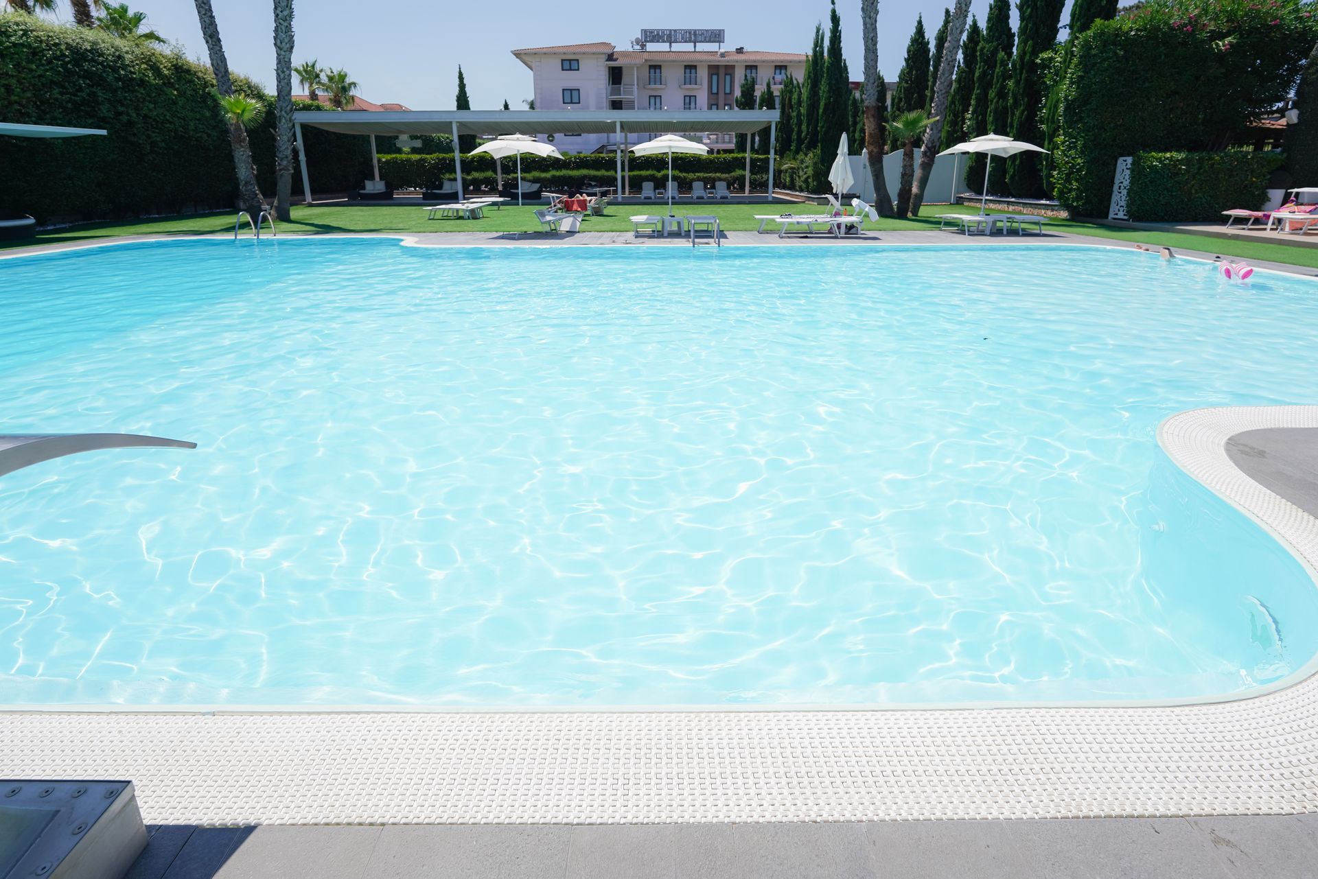 Piscina all'aperto soleggiata con acqua cristallina, lettini prendisole e un edificio sullo sfondo.