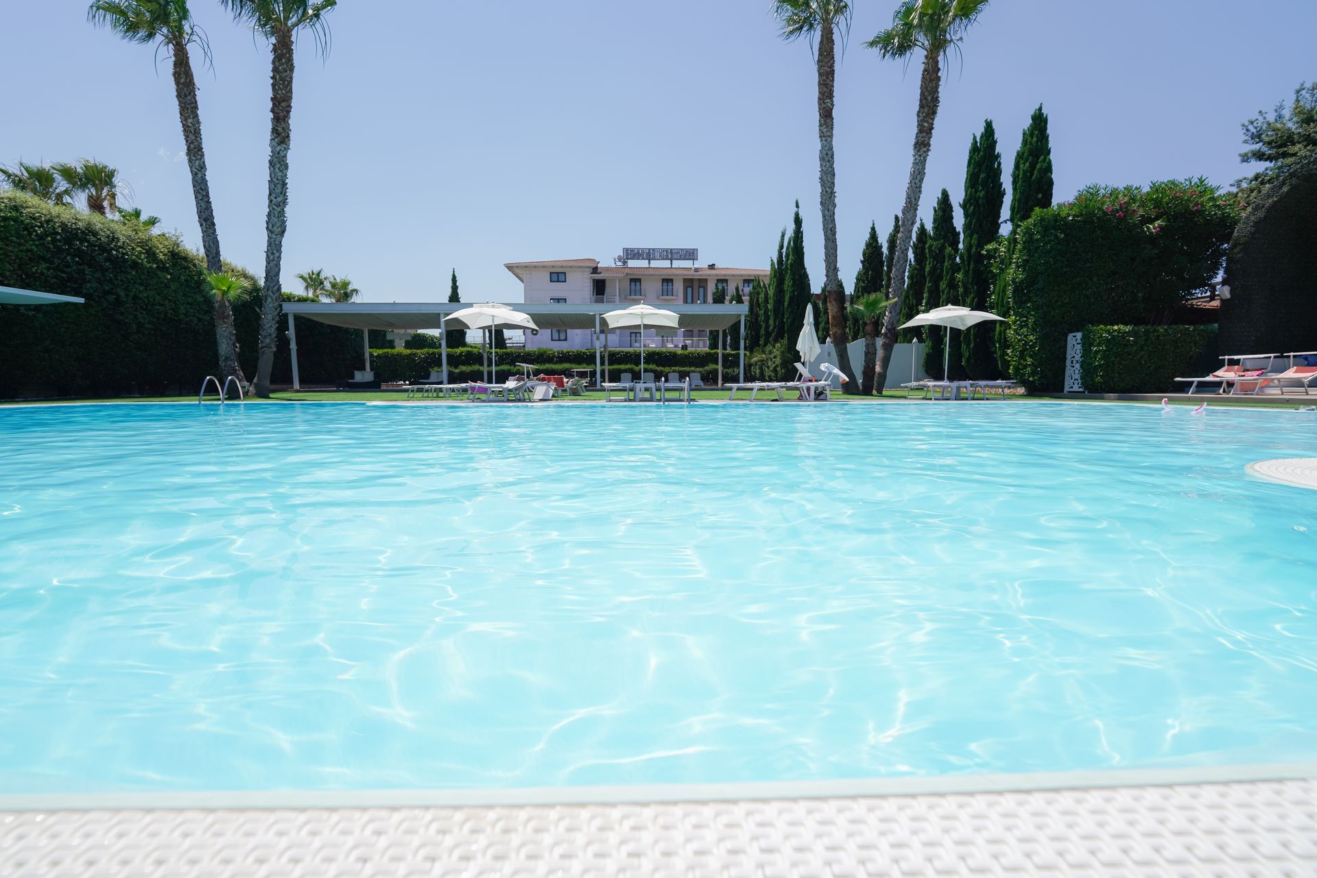 Piscina di un azzurro brillante con lettini, palme e un edificio bianco sullo sfondo.