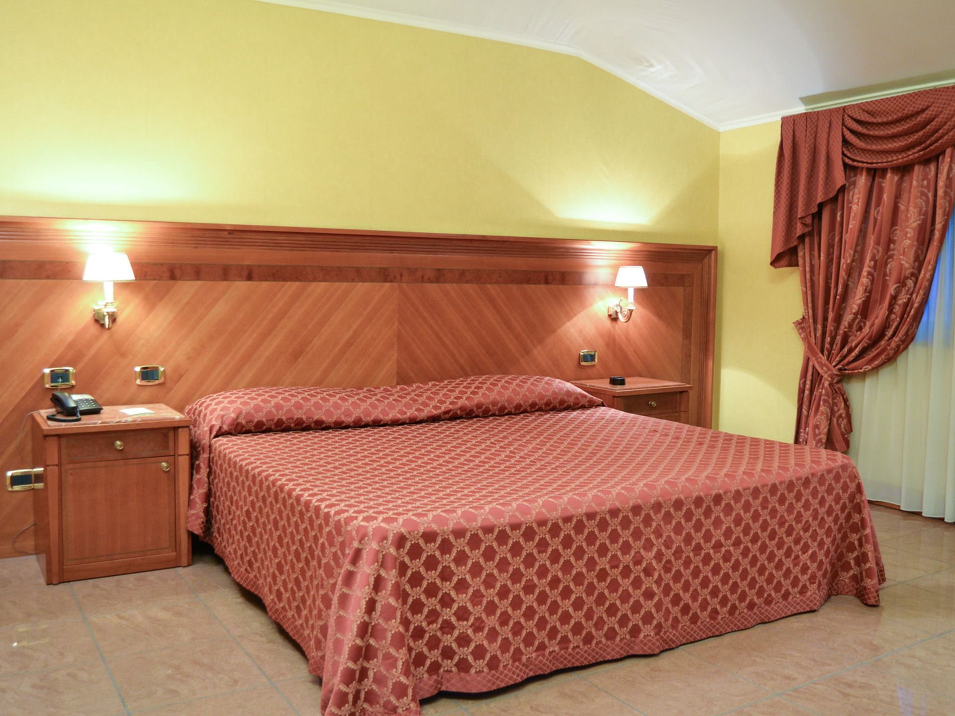Accogliente camera d'albergo con letto matrimoniale, testiera in legno, lampade da comodino e tende rosse.