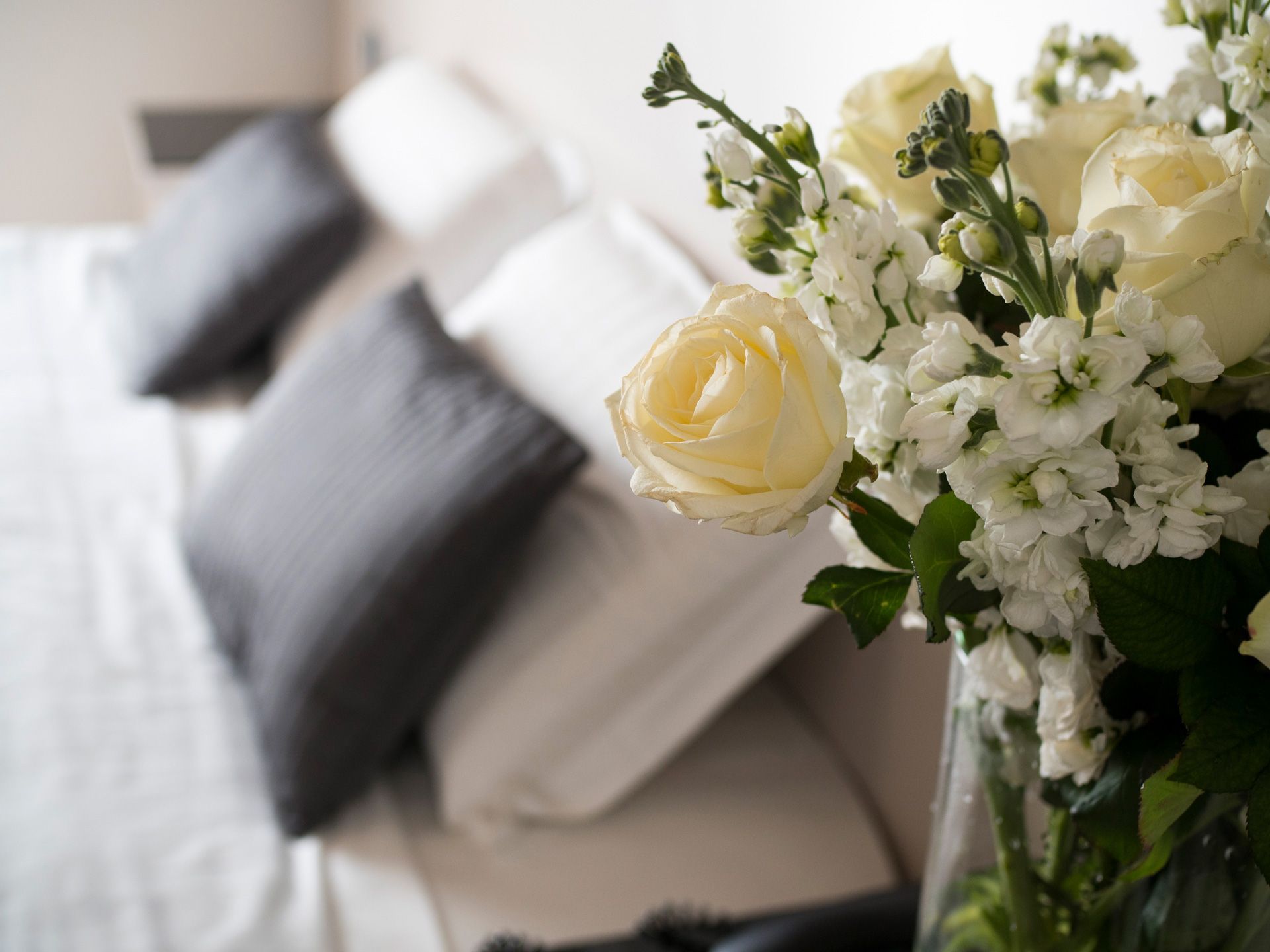 Rose bianche e fiori in un vaso accanto a un letto ben rifatto con cuscini grigi.