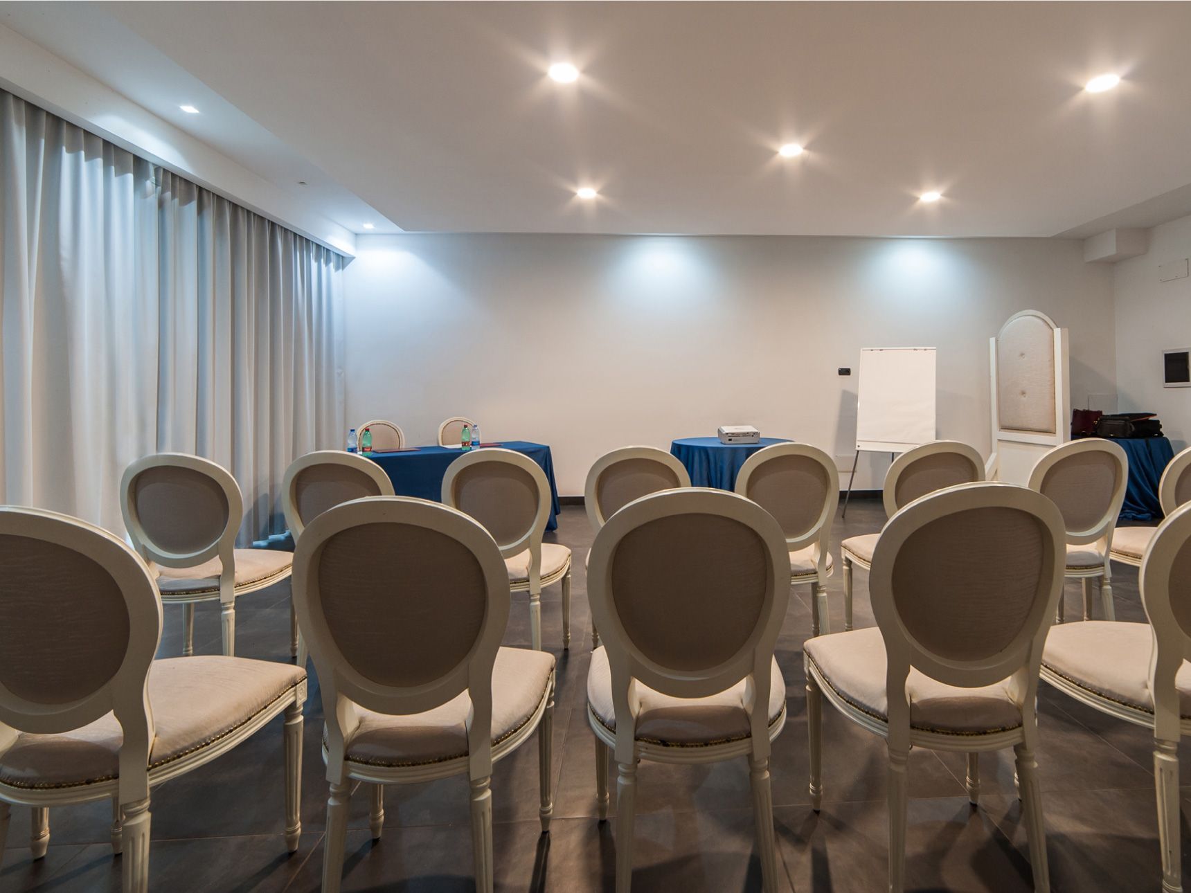 Sala conferenze vuota con file di sedie beige rivolte verso un'area di presentazione frontale.
