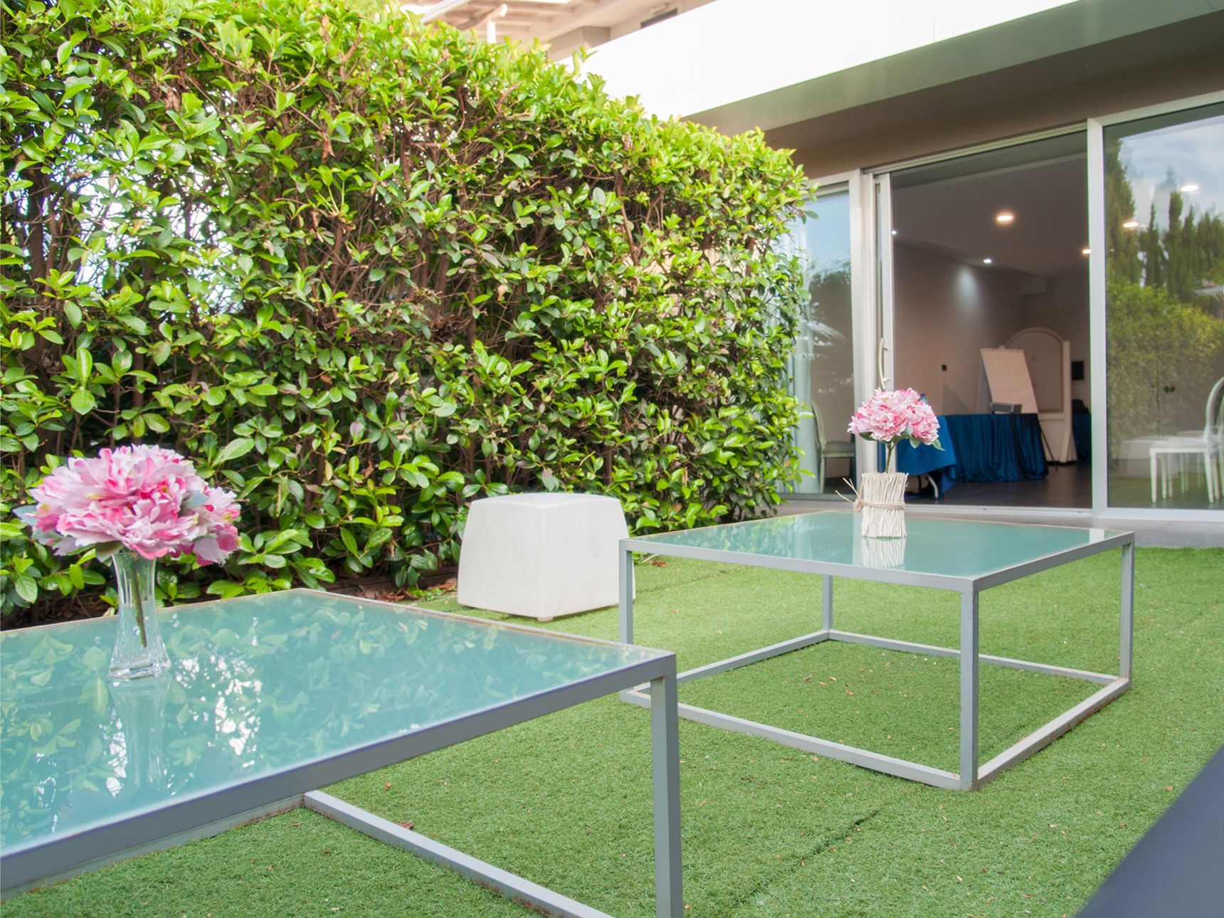 Patio con tavolini in vetro, fiori rosa, siepe verde e porte scorrevoli che conducono a una casa moderna.