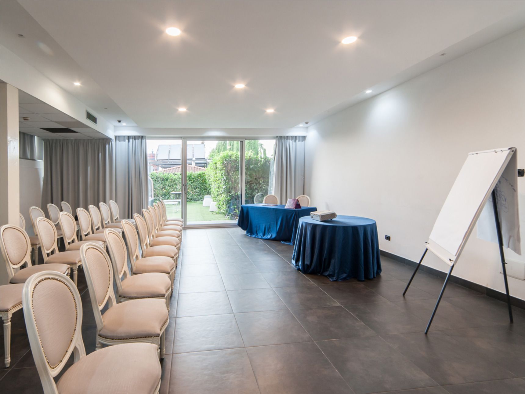 Sala conferenze vuota con file di sedie, tavoli rotondi e una lavagna a fogli mobili vicino a porte scorrevoli in vetro.