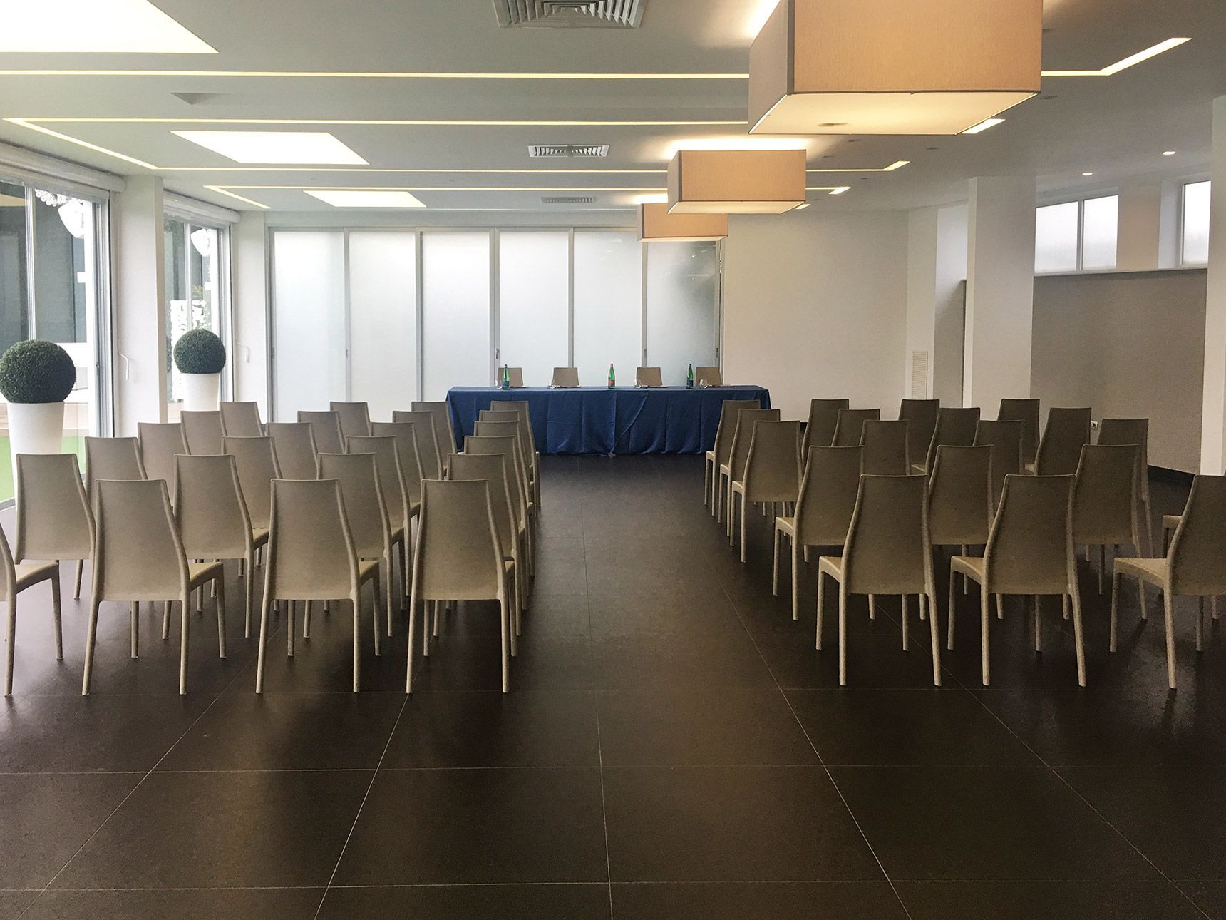 Sala conferenze vuota con file di sedie bianche disposte di fronte a un tavolo ricoperto da una tovaglia blu.