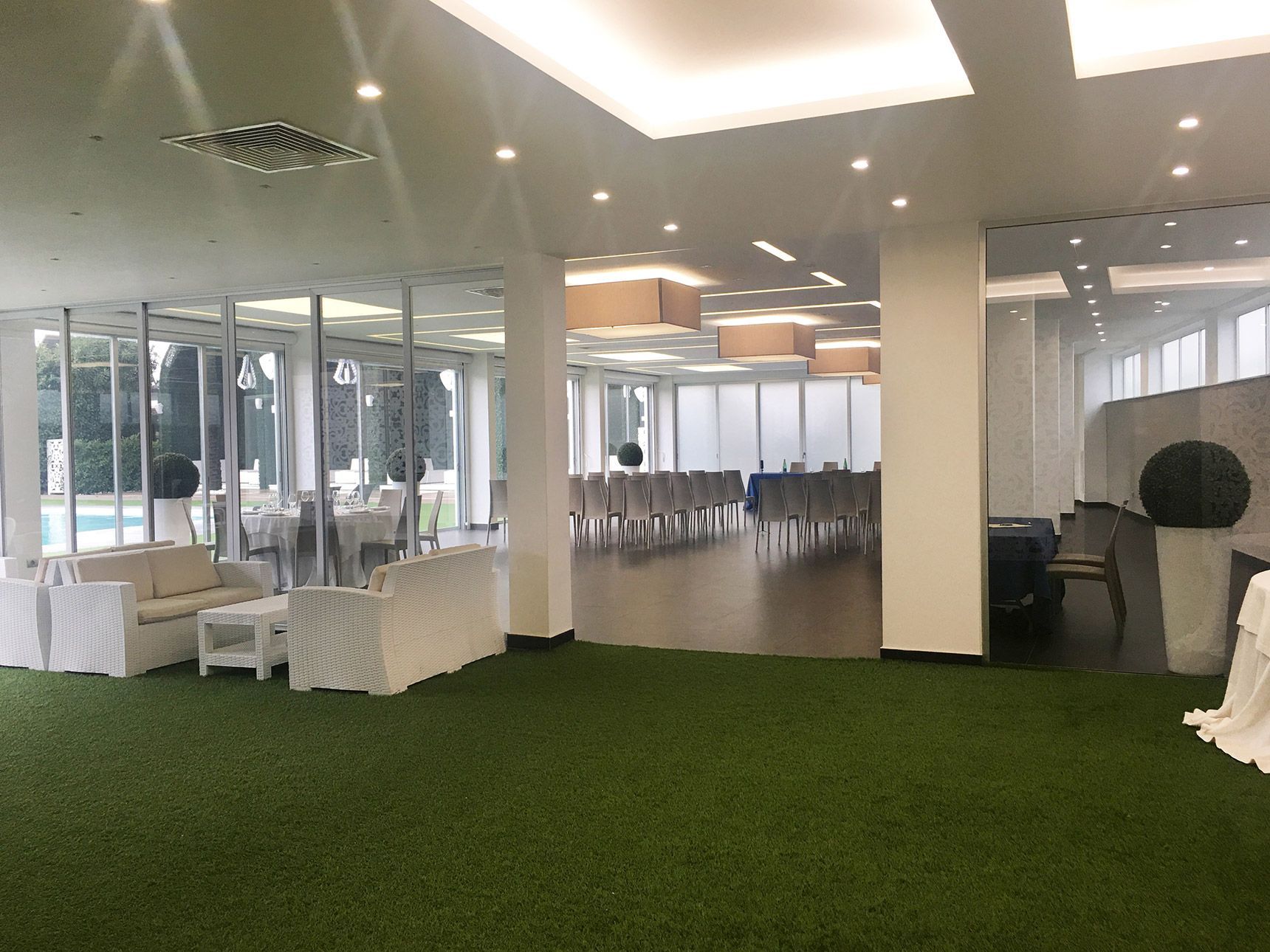 Ampia e moderna sala per banchetti con moquette verde, colonne bianche e file di sedie coperte vicino a grandi finestre.