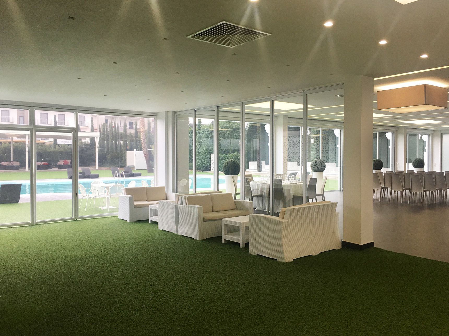 Salone moderno con pavimento in erba sintetica verde, sedute bianche, pareti in vetro e vista sulla piscina esterna.
