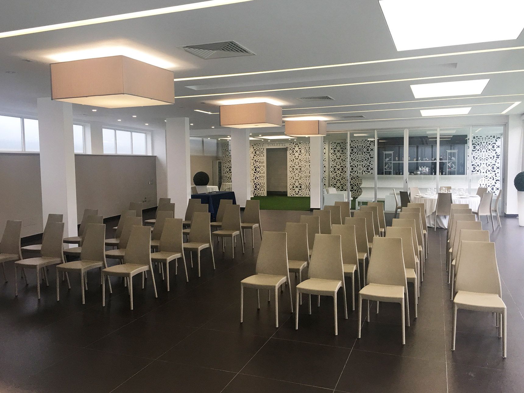 Sala conferenze moderna e vuota con file di sedie e tavoli beige sotto luci intense a soffitto.