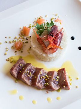 Piatto gourmet impiattato con carne scottata a fette, salmone, erbe aromatiche e salse su un piatto bianco.