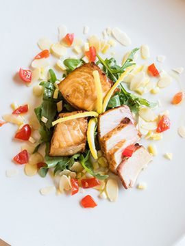 Salmone scottato servito con verdure, salsa bianca e guarnizione di verdure rosse a dadini su un piatto bianco.